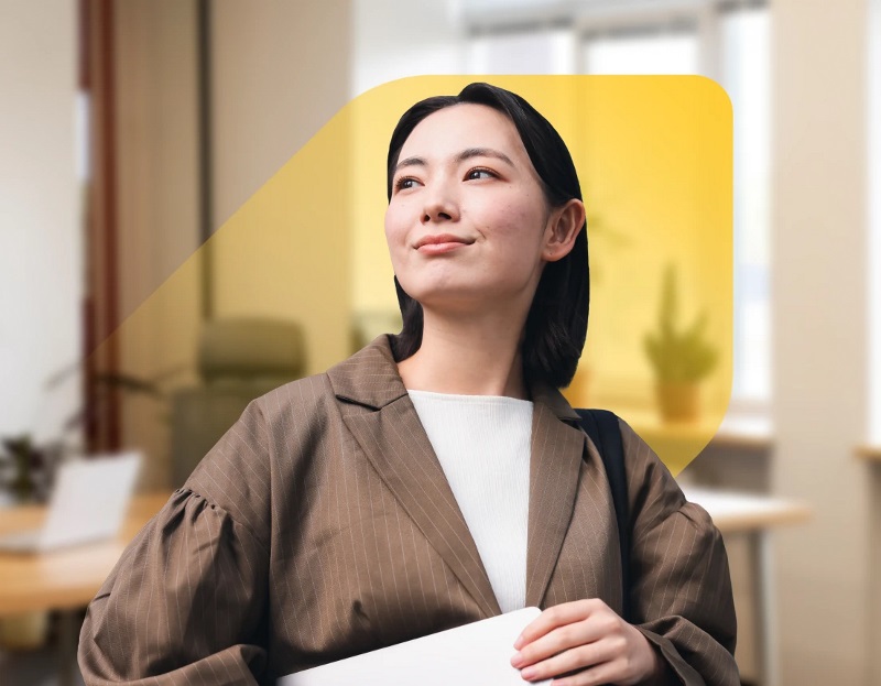 Une photo d’une femme asiatique souriant fièrement, superposée sur le losange étendu de la Banque EQ et un espace de bureau lumineux avec des plantes en pot.